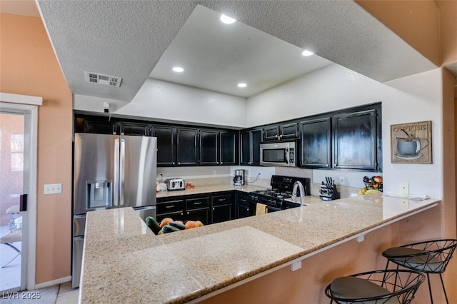 kitchen with stainless steel appliances, light stone countertops, dark cabinets, a peninsula, and a kitchen breakfast bar