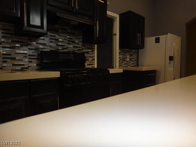 kitchen featuring dark cabinets, white refrigerator with ice dispenser, black stove, light countertops, and tasteful backsplash