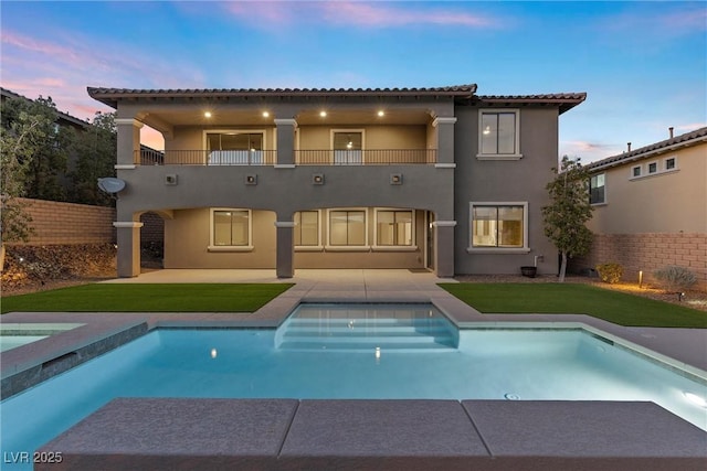 back of property at dusk featuring a fenced in pool, a balcony, fence, a patio area, and stucco siding