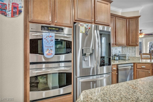 kitchen with stainless steel appliances, ornamental molding, brown cabinetry, and light stone countertops