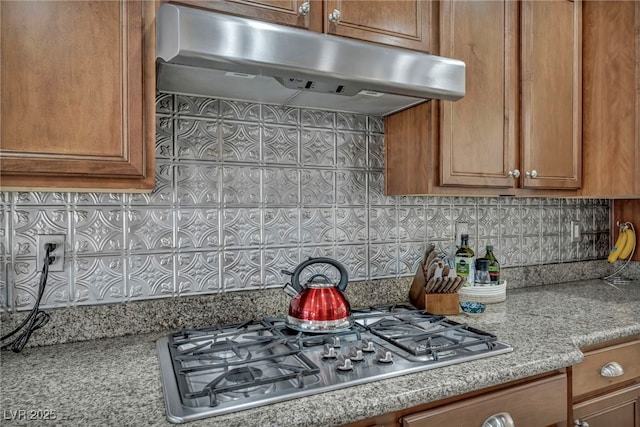 kitchen with brown cabinetry, decorative backsplash, light stone countertops, ventilation hood, and stainless steel gas cooktop