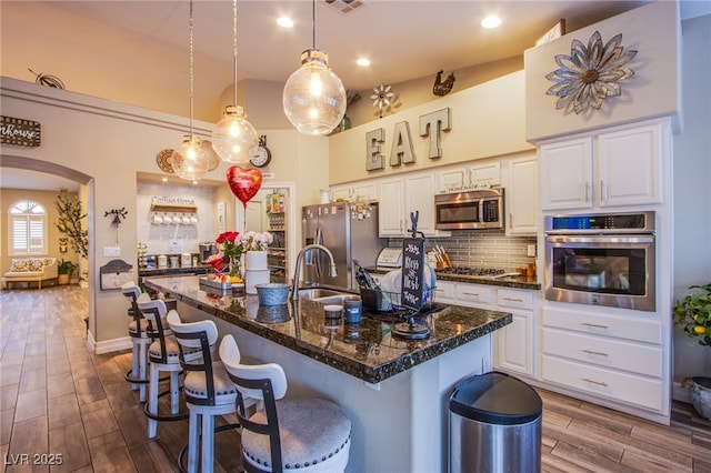 kitchen with backsplash, stainless steel appliances, a kitchen island with sink, decorative light fixtures, and white cabinetry