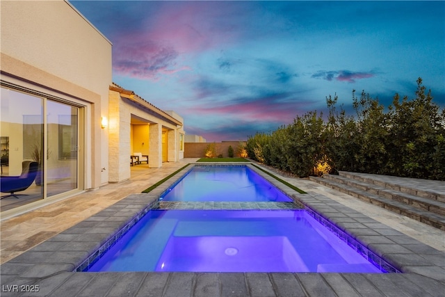 pool at dusk featuring a patio area, a pool with connected hot tub, and fence