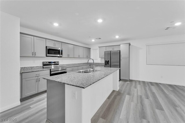 kitchen featuring light wood finished floors, an island with sink, light stone countertops, stainless steel appliances, and a sink