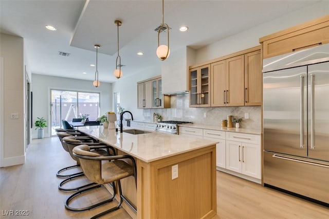 kitchen with light brown cabinets, a sink, high quality appliances, an island with sink, and glass insert cabinets