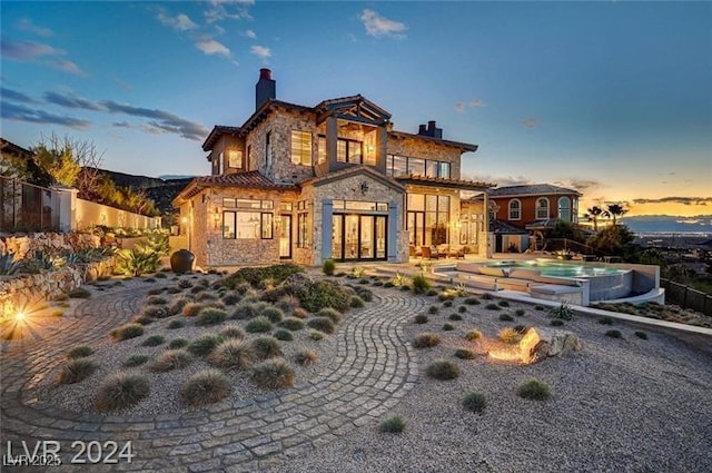 back of house at dusk featuring a hot tub, stone siding, a chimney, fence, and a patio area