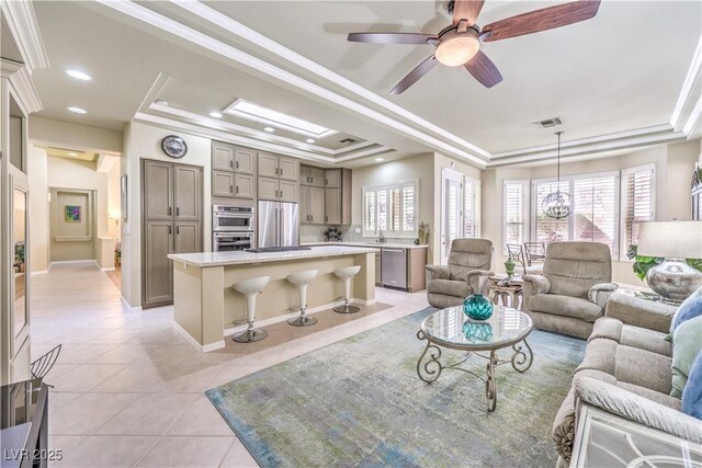 living area featuring light tile patterned floors, ceiling fan with notable chandelier, visible vents, ornamental molding, and a tray ceiling