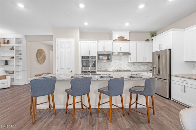 kitchen featuring a center island with sink, white cabinets, a kitchen breakfast bar, stainless steel appliances, and under cabinet range hood