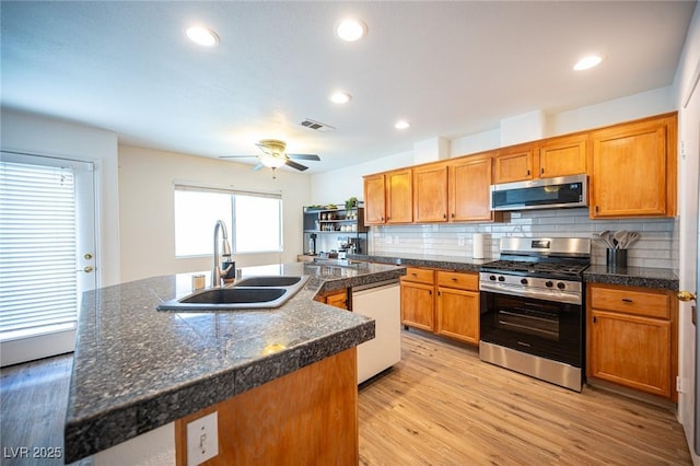 kitchen with dark countertops, brown cabinets, a kitchen island with sink, stainless steel appliances, and a sink