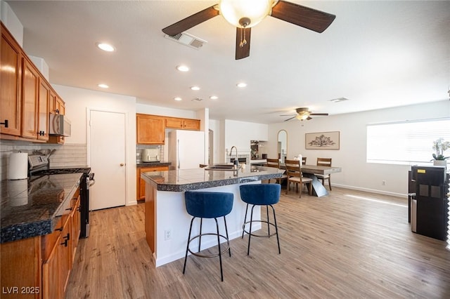 kitchen featuring stainless steel gas range oven, tile counters, brown cabinetry, an island with sink, and freestanding refrigerator