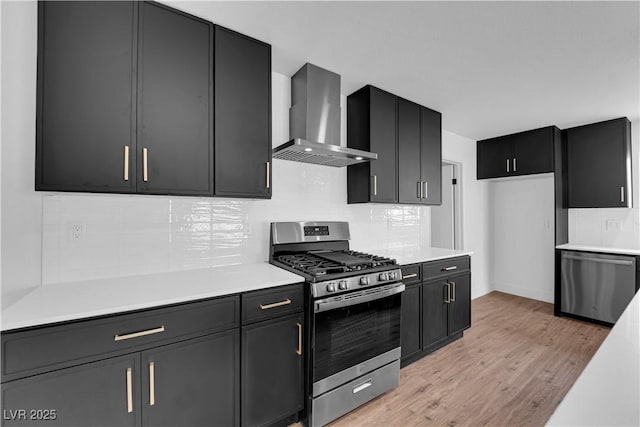 kitchen with stainless steel appliances, light countertops, wall chimney range hood, light wood-type flooring, and dark cabinetry