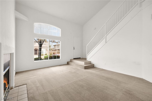 unfurnished living room featuring light carpet, a fireplace with flush hearth, stairs, and a towering ceiling