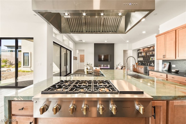 kitchen featuring decorative backsplash, light stone counters, wall chimney range hood, stainless steel gas cooktop, and a sink