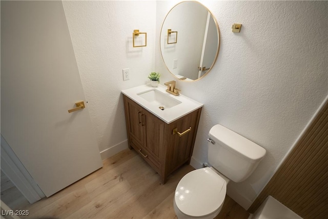 half bathroom featuring a textured wall, toilet, vanity, wood finished floors, and baseboards