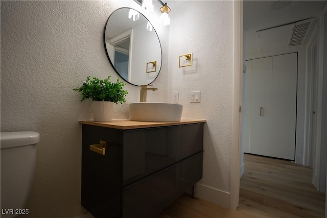 half bath featuring baseboards, visible vents, a textured wall, wood finished floors, and vanity