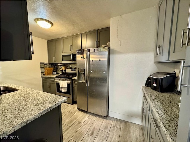 kitchen featuring wood finish floors, backsplash, gray cabinetry, appliances with stainless steel finishes, and light stone countertops