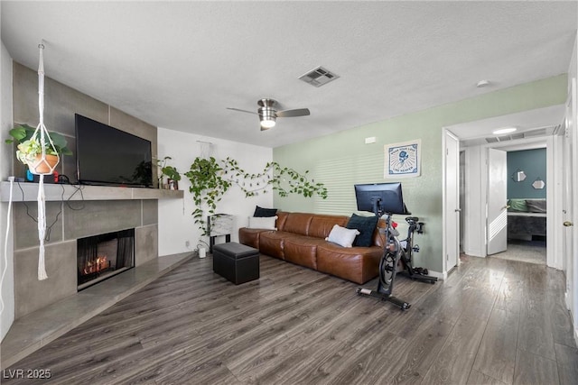 living room featuring a warm lit fireplace, a textured ceiling, wood finished floors, visible vents, and a ceiling fan