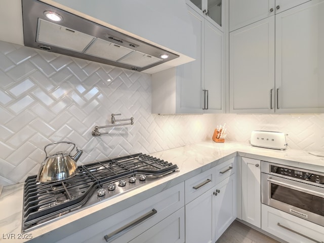 kitchen featuring appliances with stainless steel finishes, exhaust hood, glass insert cabinets, and tasteful backsplash