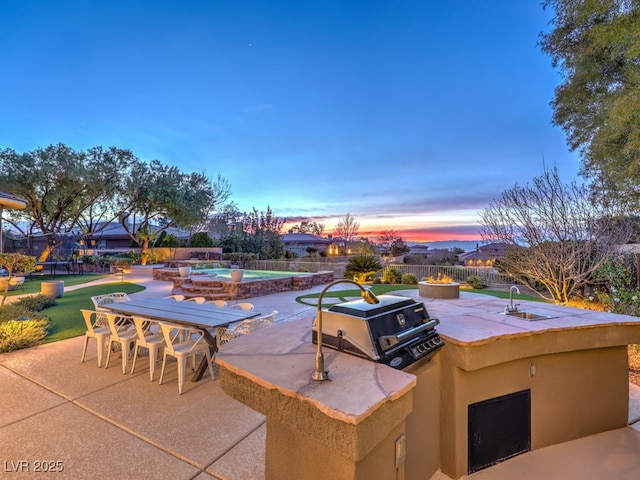 patio terrace at dusk with an outdoor kitchen, grilling area, fence, outdoor dining area, and a swimming pool