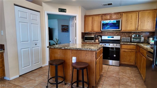 kitchen featuring a center island, brown cabinets, tasteful backsplash, appliances with stainless steel finishes, and dark stone countertops