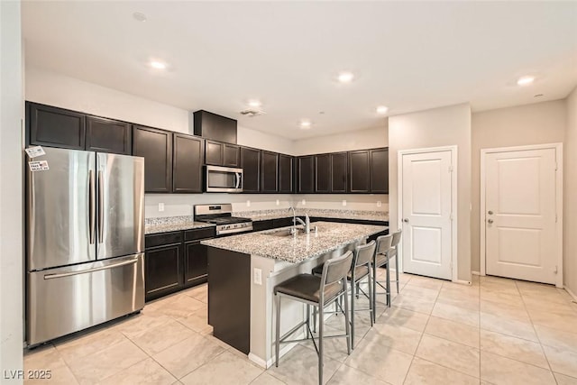 kitchen featuring a center island with sink, appliances with stainless steel finishes, a breakfast bar, light stone countertops, and a sink