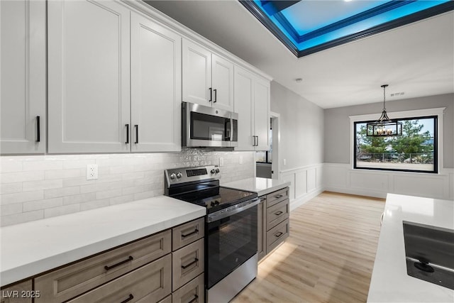 kitchen featuring a wainscoted wall, light wood finished floors, stainless steel appliances, white cabinets, and decorative light fixtures