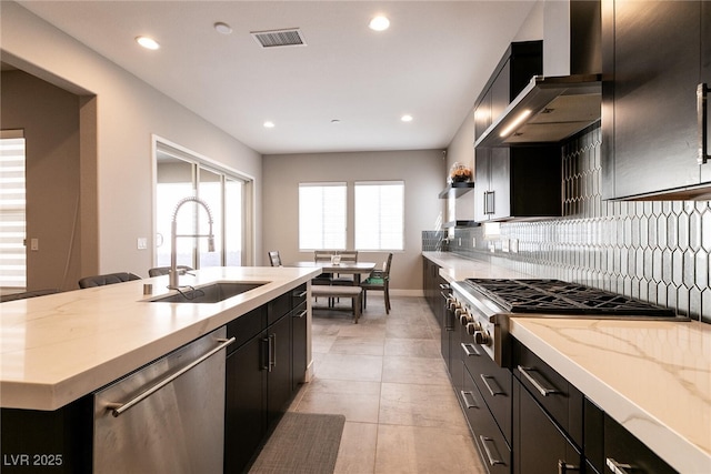 kitchen with an island with sink, dark cabinets, stainless steel appliances, wall chimney range hood, and a sink