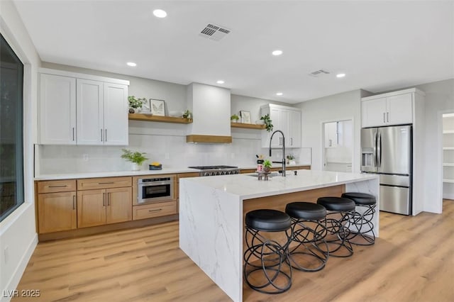 kitchen featuring an island with sink, stainless steel appliances, white cabinetry, open shelves, and a sink