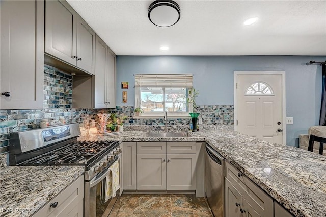 kitchen with light stone counters, gray cabinetry, stainless steel appliances, a sink, and backsplash