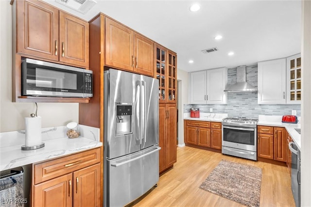 kitchen with wall chimney exhaust hood, appliances with stainless steel finishes, brown cabinetry, and glass insert cabinets