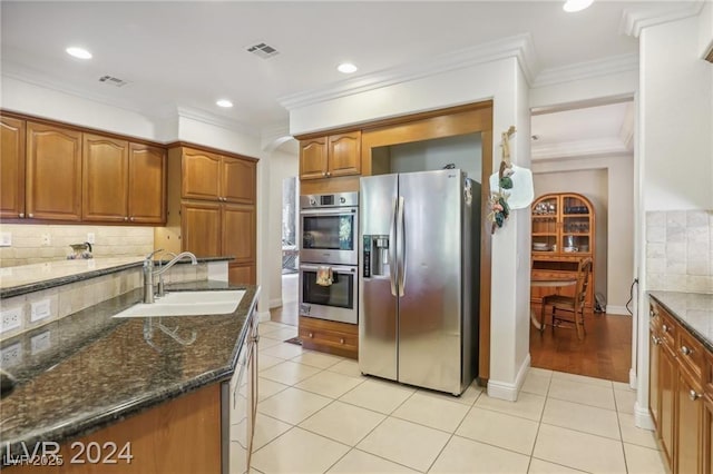 kitchen with brown cabinetry, dark stone counters, ornamental molding, stainless steel appliances, and a sink