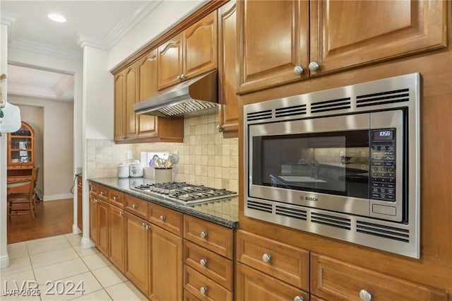 kitchen with under cabinet range hood, stainless steel appliances, ornamental molding, backsplash, and dark stone countertops