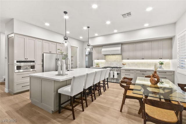 kitchen featuring premium appliances, premium range hood, a center island with sink, and gray cabinetry