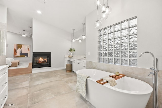 bathroom with ceiling fan, a multi sided fireplace, vanity, a freestanding bath, and vaulted ceiling