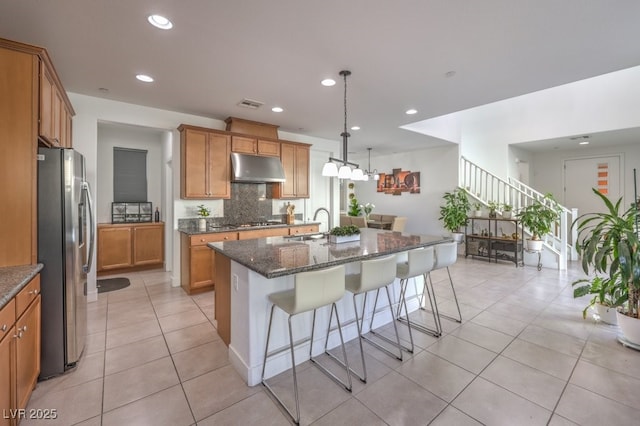 kitchen with appliances with stainless steel finishes, dark stone countertops, a kitchen island with sink, under cabinet range hood, and pendant lighting