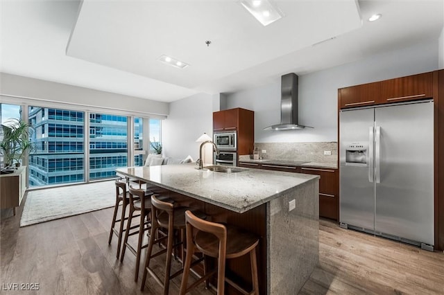 kitchen featuring a breakfast bar area, stainless steel appliances, backsplash, a kitchen island with sink, and wall chimney range hood