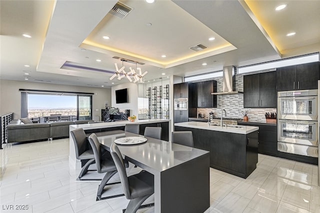 kitchen featuring wall chimney exhaust hood, a tray ceiling, an island with sink, and visible vents