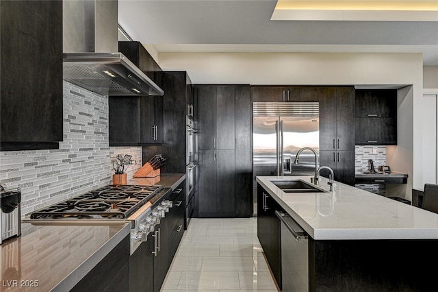 kitchen featuring a center island with sink, backsplash, appliances with stainless steel finishes, a sink, and wall chimney exhaust hood