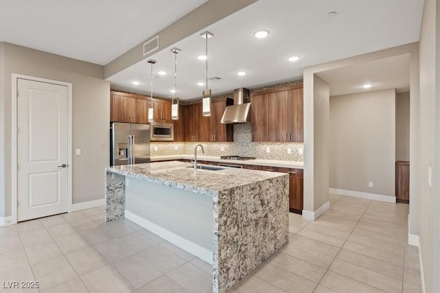 kitchen featuring decorative light fixtures, a center island with sink, appliances with stainless steel finishes, a sink, and wall chimney exhaust hood