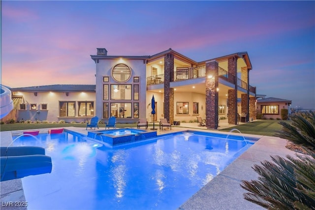 back of house at dusk featuring a balcony, a pool with connected hot tub, stucco siding, a chimney, and a patio area