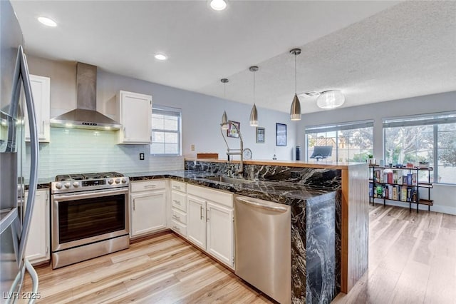 kitchen featuring white cabinets, wall chimney exhaust hood, decorative light fixtures, stainless steel appliances, and a sink