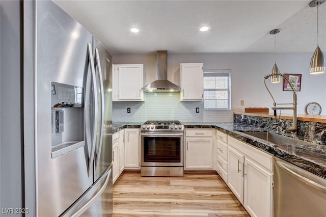 kitchen with stainless steel appliances, a sink, white cabinets, hanging light fixtures, and wall chimney range hood