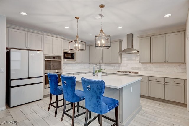 kitchen with light countertops, hanging light fixtures, appliances with stainless steel finishes, a kitchen island with sink, and wall chimney range hood