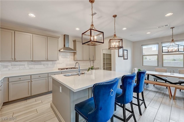 kitchen with visible vents, an island with sink, light countertops, wall chimney range hood, and a sink