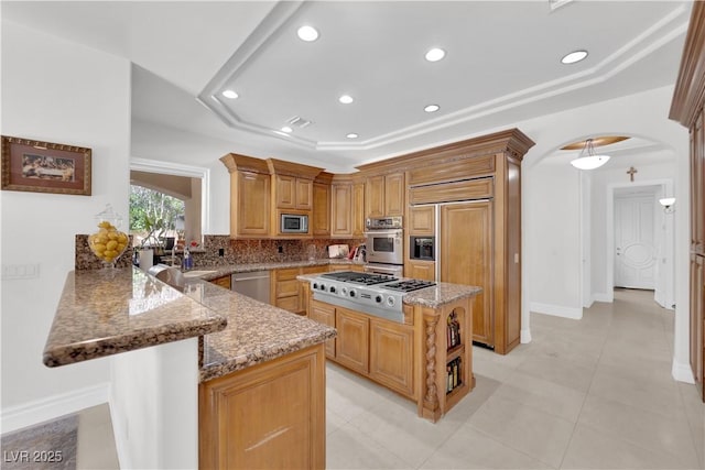 kitchen featuring arched walkways, light stone counters, built in appliances, a peninsula, and a tray ceiling