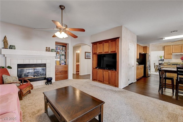 living area featuring visible vents, arched walkways, a ceiling fan, a tiled fireplace, and dark carpet