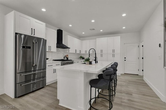 kitchen with high end refrigerator, light wood-style floors, light stone counters, wall chimney range hood, and a sink