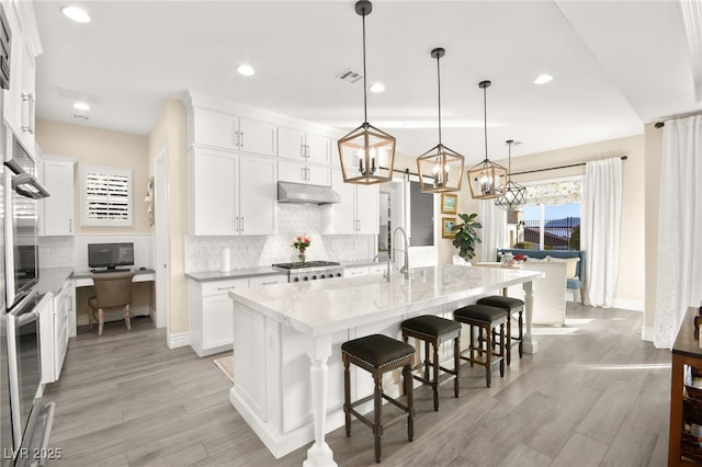 kitchen featuring light wood-style flooring, under cabinet range hood, a kitchen bar, stainless steel oven, and tasteful backsplash