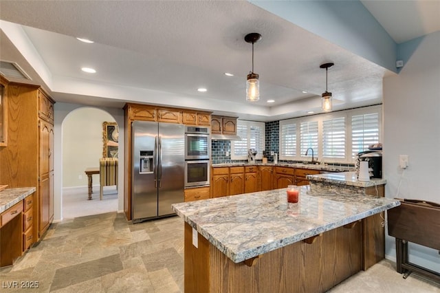 kitchen with arched walkways, brown cabinets, a raised ceiling, appliances with stainless steel finishes, and a peninsula