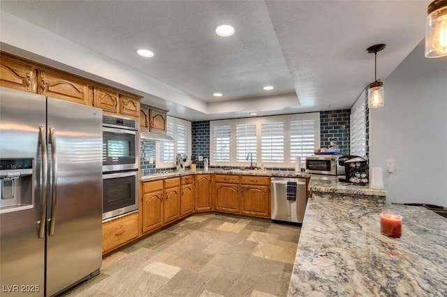 kitchen with recessed lighting, appliances with stainless steel finishes, brown cabinets, light stone countertops, and a raised ceiling